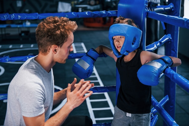 Der junge Boxtrainer hilft dem kleinen Jungen in Schutzkleidung auf dem Ring zwischen den Runden.