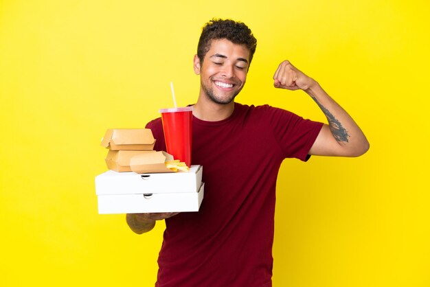 Der junge Brasilianer, der Pizzen und Burger hält, isoliert den Hintergrund und macht eine starke Geste