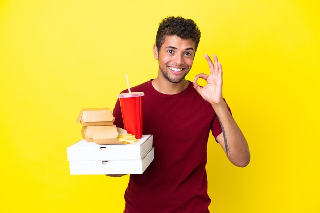 Der junge Brasilianer, der Pizzen und Burger hält, isoliert den Hintergrund und zeigt ein Ok-Zeichen mit den Fingern