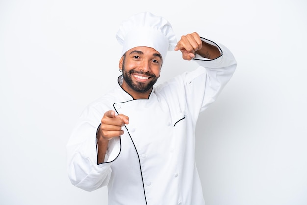 Der junge brasilianische Koch, der auf weißem Hintergrund isoliert ist, zeigt mit dem Finger auf Sie, während er lächelt