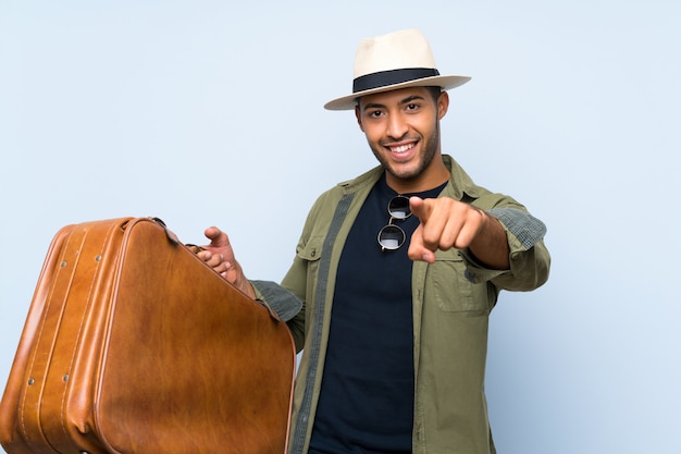 Der junge gutaussehende Mann, der einen Weinleseaktenkoffer über lokalisierter blauer Wand hält, zeigt Finger auf Sie mit einem überzeugten Ausdruck