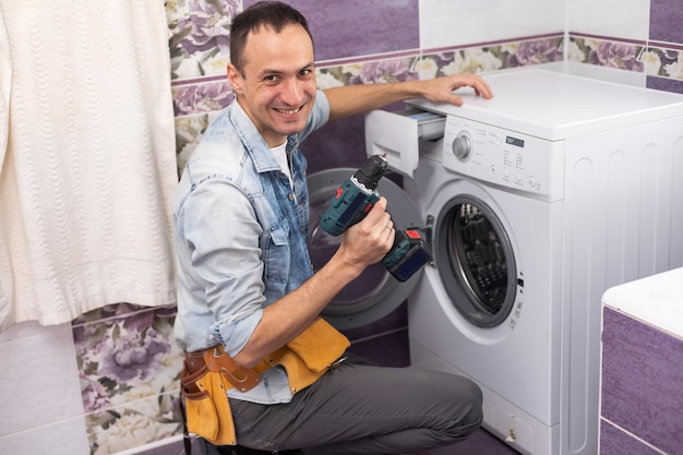 Der junge gutaussehende Mechaniker im Arbeiteranzug mit der professionellen Werkzeugkiste repariert die Waschmaschine im Badezimmer