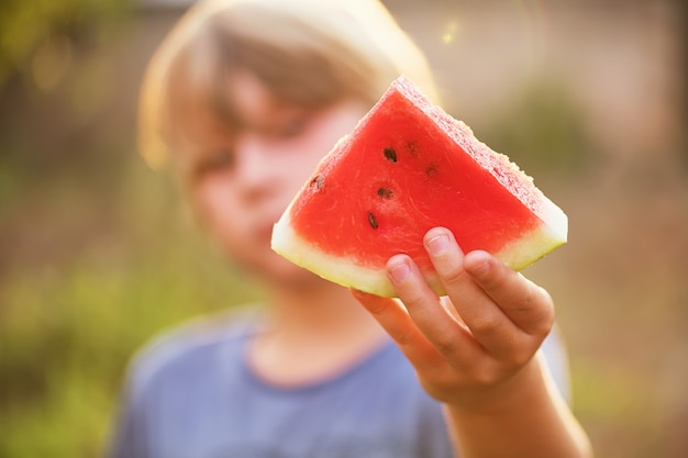 Der Junge hält ein Stück Wassermelone in der Hand und streckt es aus, selektiver Fokus, Linseneffekt. Sommerkonzept, frisch und fröhlich.