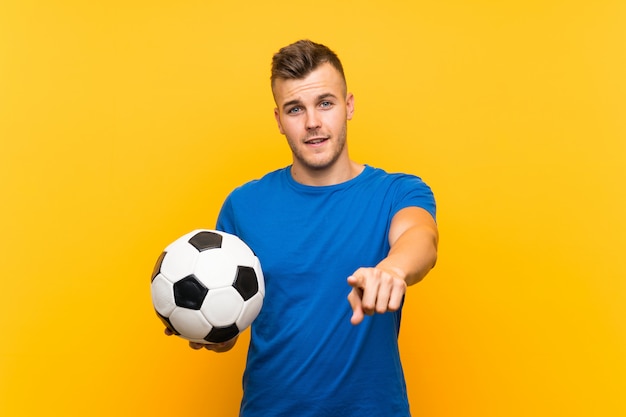 Der junge hübsche blonde Mann, der einen Fußball über lokalisierter gelber Wand hält, zeigt Finger auf Sie mit einem überzeugten Ausdruck