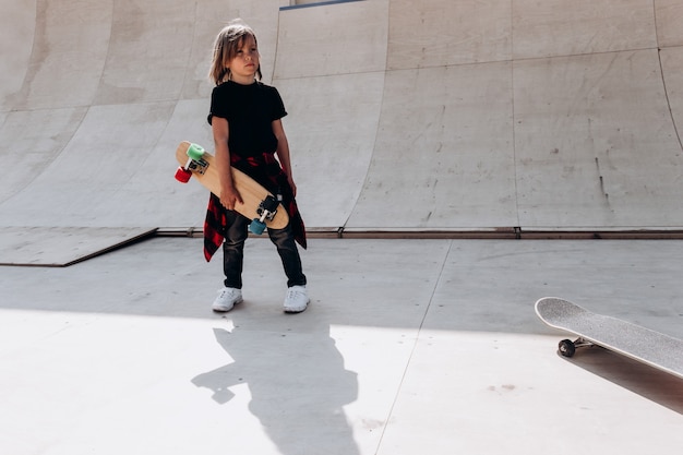 Der Junge in Freizeitkleidung mit Skateboard in der Hand steht am sonnigen Tag draußen in einem Skatepark neben der Rutsche.