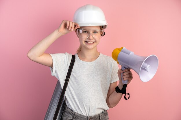 Der junge Ingenieur ist ein Mädchen an einer Wand einer rosa Wand, das einen Helm, eine Brille und einen Lautsprecher in den Händen trägt