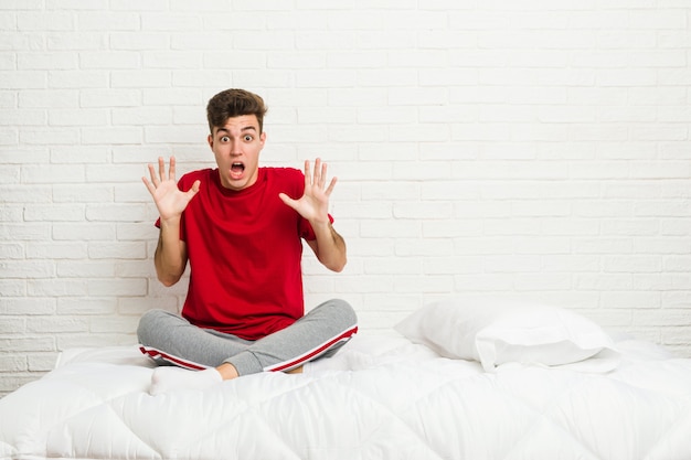 Der junge Jugendlichstudentenmann auf dem Bett einen Sieg oder einen Erfolg feiernd, ist er überrascht und entsetzt.