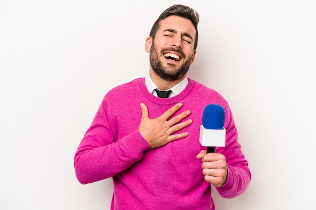 Der junge kaukasische Fernsehmoderator, der auf weißem Hintergrund isoliert ist, lacht laut und hält die Hand auf der Brust