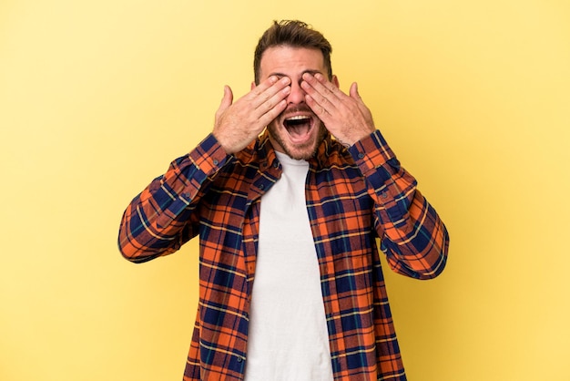 Der junge kaukasische Mann, der auf gelbem Hintergrund isoliert ist, bedeckt die Augen mit den Händen, lächelt breit und wartet auf eine Überraschung.