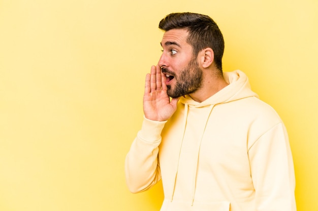 Der junge kaukasische Mann, der auf gelbem Hintergrund isoliert ist, sagt eine geheime heiße Bremsnachricht und schaut zur Seite