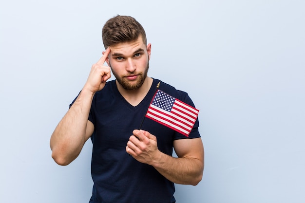 Der junge kaukasische Mann, der eine Flagge Vereinigter Staaten zeigt seinen Tempel mit dem Finger hält und denkt, konzentrierte sich auf eine Aufgabe.