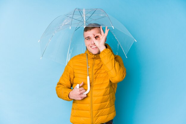 Der junge kaukasische Mann, der einen Regenschirm hält, regte das Halten der okaygeste auf Auge auf.