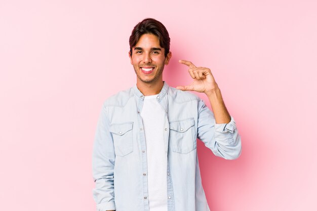 Der junge kaukasische Mann, der in einer rosa Wand aufwirft, lokalisierte das Halten etwas wenig mit den Zeigefingern, lächelte und überzeugte.