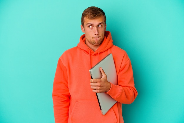 Der junge kaukasische Student, der einen Laptop hält, der auf der blauen Wand lokalisiert verwirrt ist, fühlt sich zweifelhaft und unsicher.