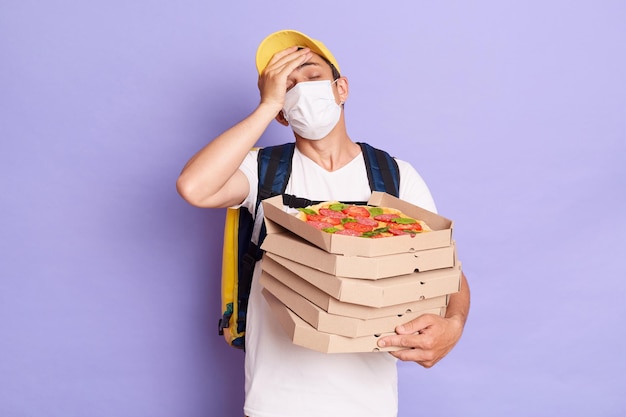 Der junge Lieferbote mit T-Shirt und Schutzmaske, der Pizzakartons in den Händen hält, vergaß die Adresse des Kunden, die eine Facelpalm-Geste zeigt, die isoliert vor violettem Hintergrund posiert