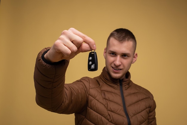 Der junge Mann der 20er Jahre in einer braunen Jacke auf gelbem Hintergrund, der in der Hand hält, gibt einen neuen Autoschlüssel