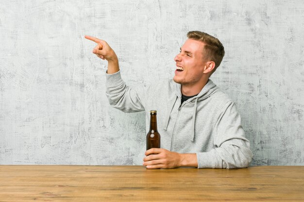 Der junge Mann, der ein Bier auf einer Tabelle trinkt, regte das Zeigen mit den Zeigefingern weg auf.