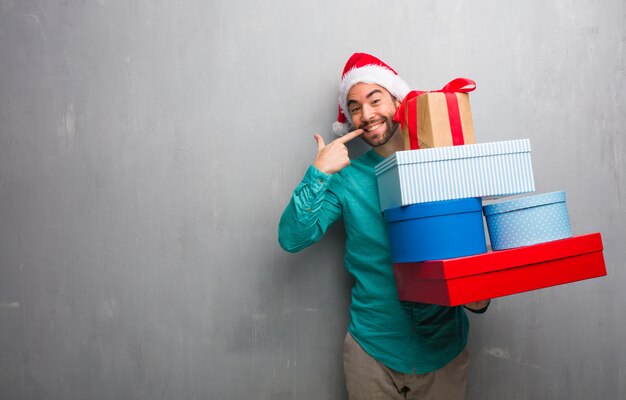 Der junge Mann, der einen Sankt-Hut hält Geschenke trägt, lächelt und zeigt Mund