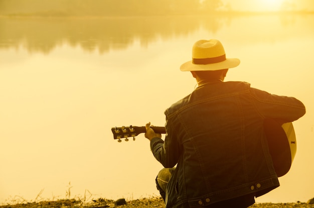 Der junge Mann, der Gitarre neben einem See mit Sonnenuntergang spielt