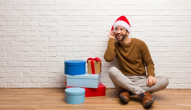 Der junge Mann, der mit den Geschenken sitzt, die Weihnachten feiern, versuchen zum Hören eines Klatsches