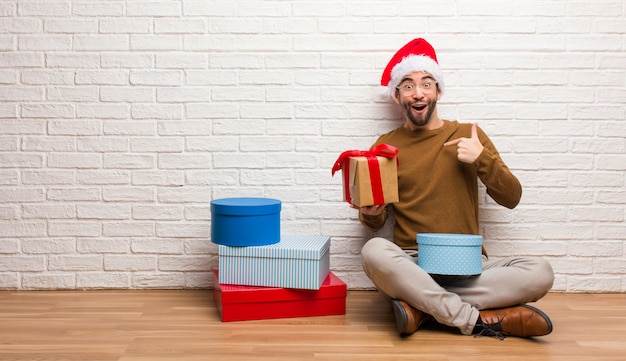 Der junge Mann, der mit den Geschenken sitzt, die Weihnachten überrascht feiern, fühlt sich erfolgreich und wohlhabend