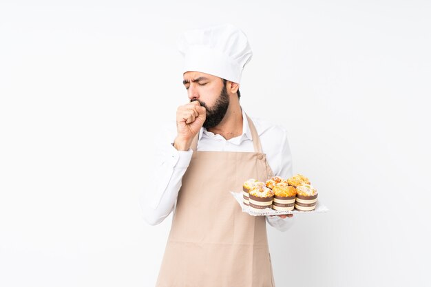 Der junge Mann, der Muffinkuchen über lokalisierter weißer Wand hält, leidet mit Husten und fühlt sich schlecht