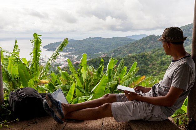 Der junge Mann, der unter Verwendung des Laptops gegen Landschaft sitzt, gestalten Wald im Sonnenuntergang landschaftlich