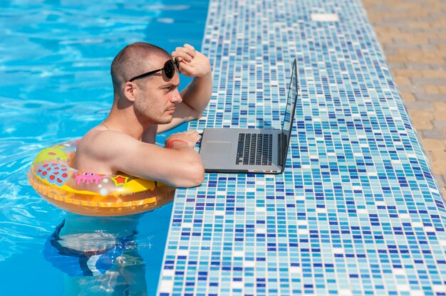 Der junge Mann in den Sommerferien mit dem Laptop, Geschäft online. Entfernung Home Office im Schwimmbad im Gummiring auf Quarantäne