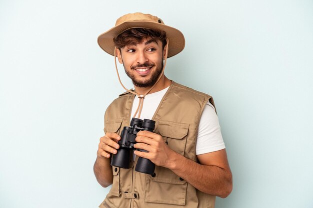 Der junge Mann mit gemischter Rasse, der ein Fernglas isoliert auf blauem Hintergrund hält, sieht beiseite lächelnd, fröhlich und angenehm aus.