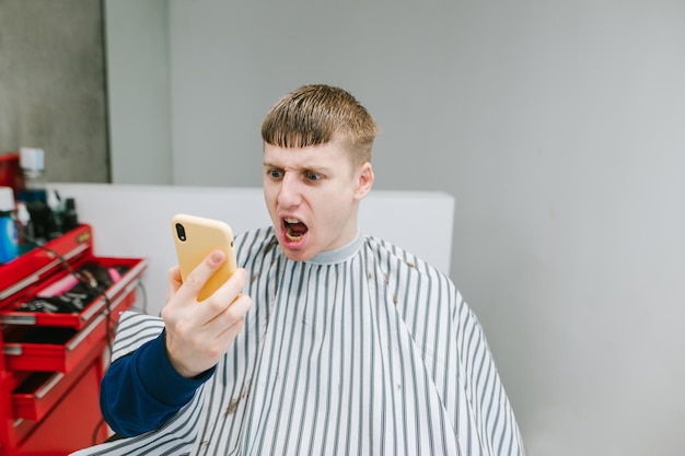 Der junge Mann sitzt mit einem Smartphone in der Hand auf einem Friseurstuhl und schaut auf einen Smartphone-Bildschirm