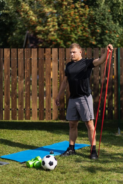 Der junge Mann treibt am Sommertag zu Hause im Hinterhof Sport. Junger Sportler mit blonden Haaren schüttelt Hände mit Sportgummi auf Matte, es gibt einen Ball, Dumbeels.