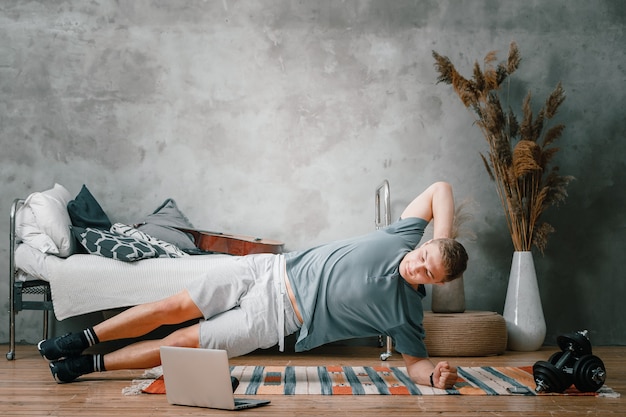 Der junge Mann treibt zu Hause Sport und trainiert online. Der Athlet hält die Planke auf der Seite, schaut auf einem Laptop im Schlafzimmer auf die Zeit, im Hintergrund ein Bett, eine Vase, einen Teppich.