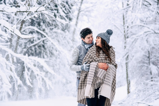 Der junge Mann umarmt zärtlich seine schöne schwangere Frau, die mit einer warmen Decke mit Schneefall bedeckt ist