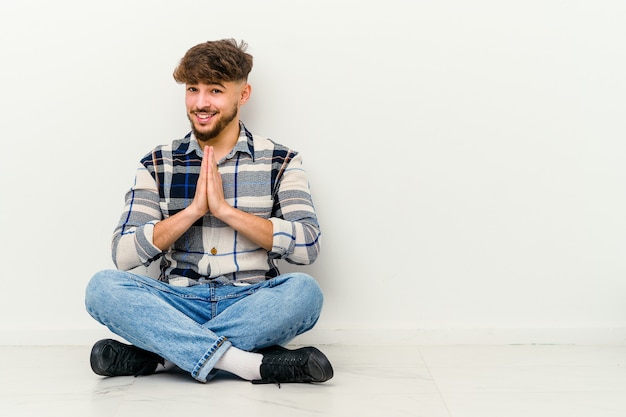 Der junge marokkanische Mann, der auf dem Boden sitzt, isoliert auf weißen Händchenhalten im Gebet nahe Mund, fühlt sich zuversichtlich.