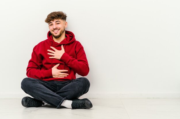 Der junge marokkanische Mann, der isoliert auf Weiß auf dem Boden sitzt, lacht glücklich und hat Spaß daran, Hände auf dem Bauch zu halten.