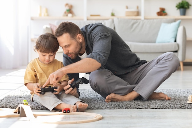 Der junge Papa erklärt dem kleinen Sohn, wie man mit dem Joystick spielt, und bringt seinem Kind bei, Videospiele zu spielen, die gemeinsam Spaß haben