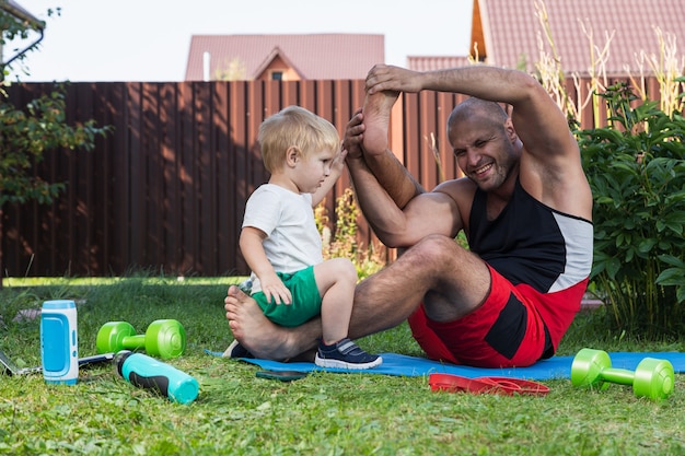 Der junge Papa-Sportler mit seinem kleinen fröhlichen Sohn treibt Sport, macht Yoga, wirft an einem warmen Tag im Garten in der Nähe des Hauses sein Bein über den Kopf auf die Matte