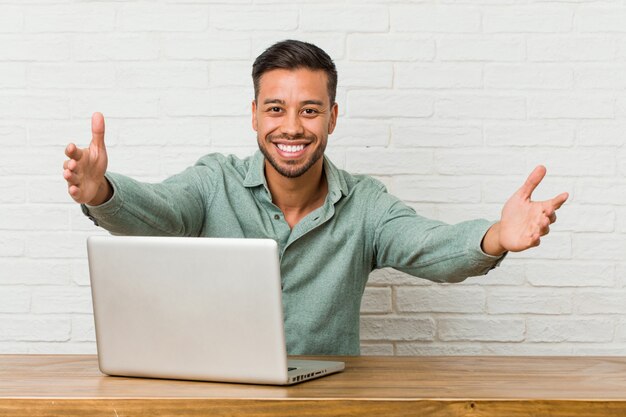 Der junge philippinische Mann, der das Arbeiten mit seinem Laptop sitzt, glaubt, eine Umarmung zu geben überzeugt