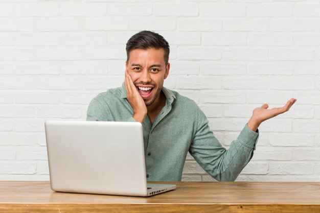 Der junge philippinische Mann, der mit seinem Laptop arbeitet, hält Kopienraum auf einer Handfläche, Hand über Wange halten. Erstaunt und entzückt.