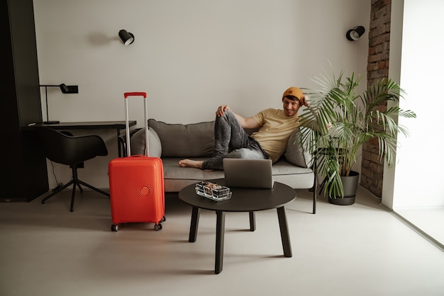 Der junge Reisende sitzt auf der Couch und sieht sich im Sommerurlaub etwas auf dem Laptop im Hotelzimmer an. Cooler Mann in Freizeitkleidung mit Gepäck.