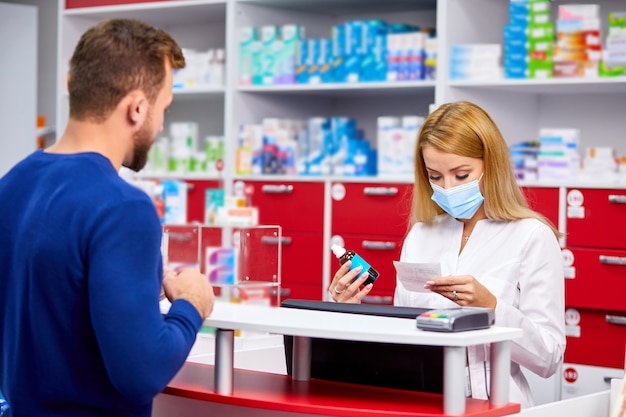 Der junge schöne kaukasische Apotheker in der medizinischen Schutzmaske arbeitet mit Kunden