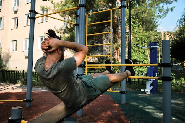 Der junge süße kaukasische Mann schüttelt seine Bauchmuskeln auf dem mit Trainingsgeräten ausgestatteten Straßensportplatz