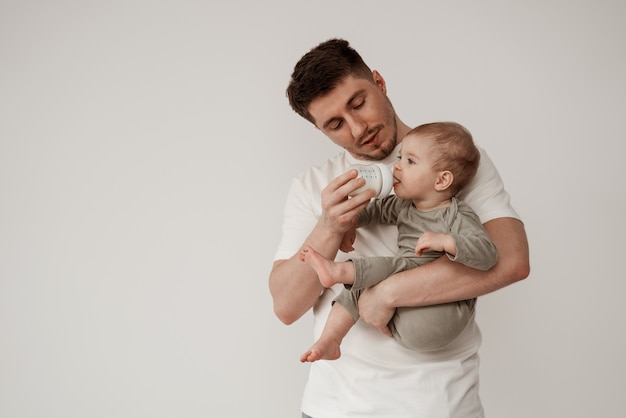 Der junge Vater füttert das Kind vorsichtig mit Muttermilch oder Säuglingsmilch. Füttern mit einer Flasche, ein Baby in den Armen halten in einem weißen Raum auf hellem Hintergrund