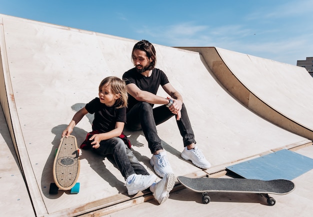 Der junge Vater und sein Sohn in der stylischen Freizeitkleidung sitzen am sonnigen warmen Tag zusammen auf der Rutsche neben den Skateboards in einem Skatepark.