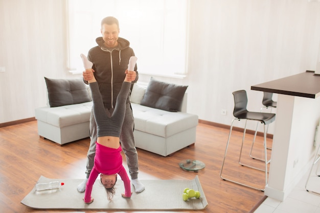 Der junge Vater und seine süße kleine Tochter trainieren zu Hause. Handstand Push-up. Nettes Kind steht auf Händen. Sie tragen Sportbekleidung und haben Übungen