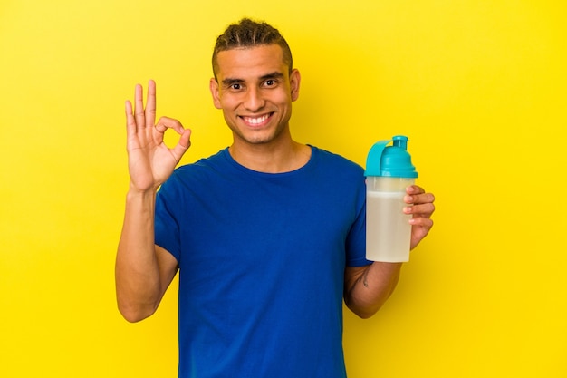 Der junge venezolanische Mann, der einen Proteinshake trinkt, isoliert auf gelbem Hintergrund, fröhlich und selbstbewusst, der eine ok Geste zeigt.