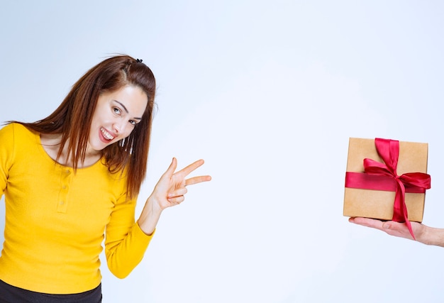 Der jungen Frau im gelben Hemd wird eine Geschenkbox aus Karton mit rotem Band und positivem Handzeichen angeboten