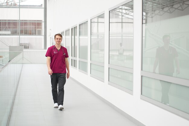 Der kaukasische junge lächelnde fröhliche Arzt hat einen Spaziergang in den Korridoren der Klinik. Person bei der Arbeit.