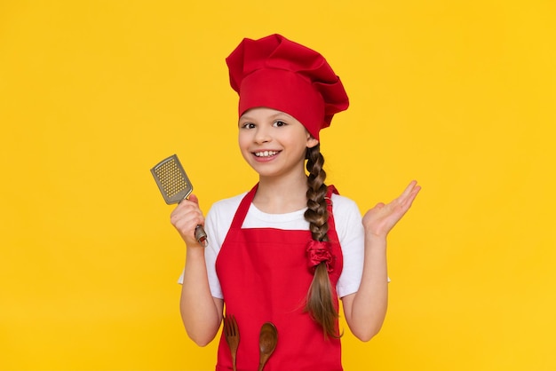 Der Kinderkoch hält eine Käsereibe und lächelt breit Ein kleines Mädchen in einem roten Hut und einer Kochschürze auf gelb isoliertem Hintergrund