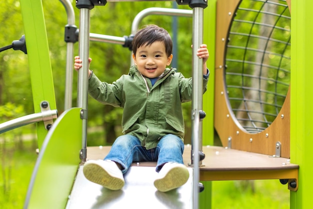 Der kleine asiatische Junge spielt im Sommer im Freien auf dem Holzspielplatz mit fröhlichem Gesicht und schaut mit einem Lächeln auf die Kamera